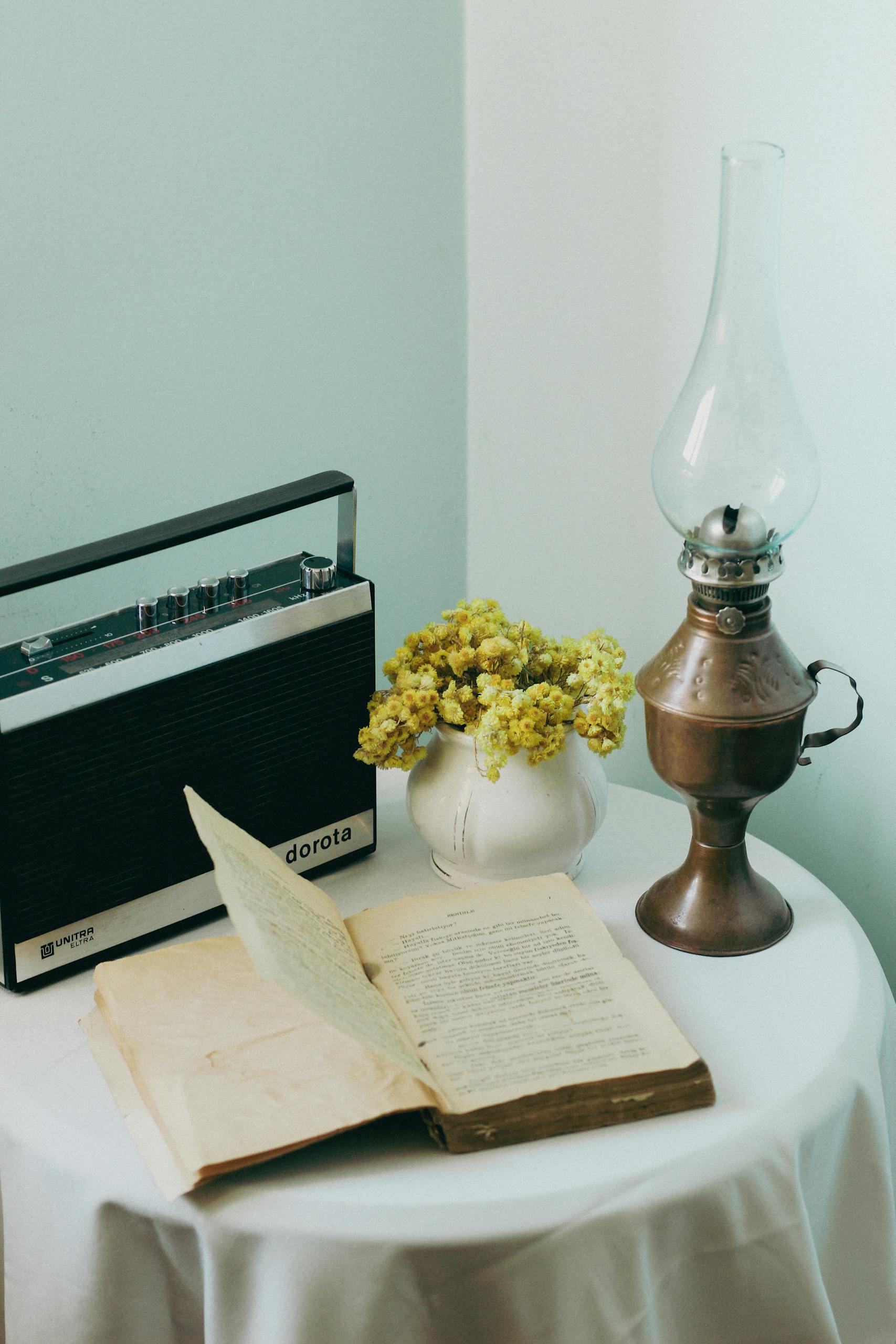 A vintage tabletop setting with a retro radio, open book, brass lamp, and flowers.