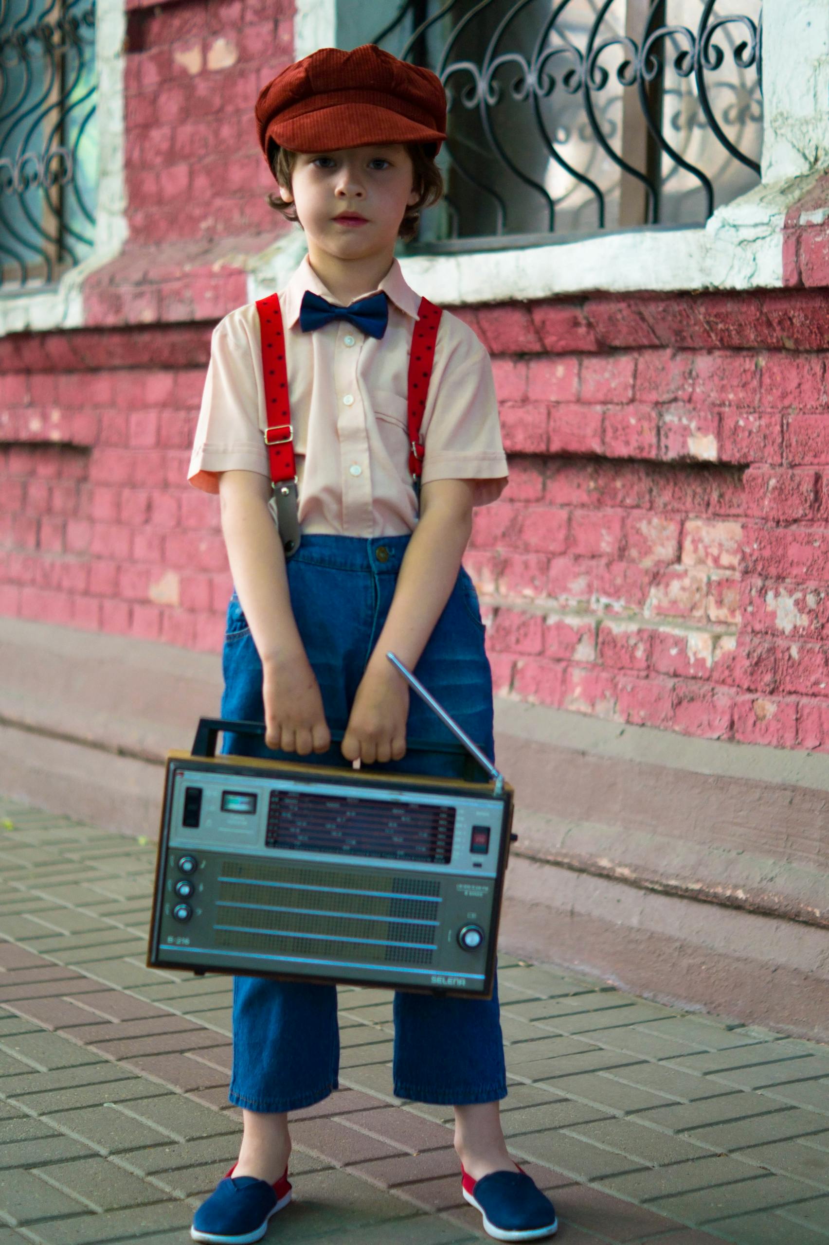 Young boy in retro fashion holding a vintage radio against a brick wall backdrop. Outdoors scene.