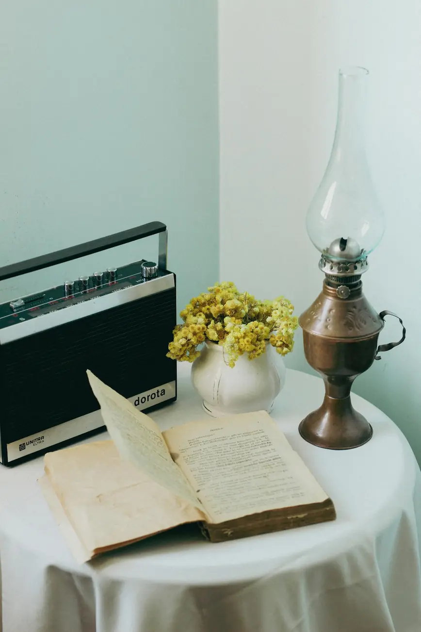 A vintage tabletop setting with a retro radio, open book, brass lamp, and flowers.