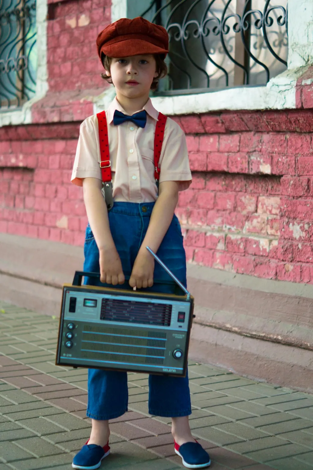 Young boy in retro fashion holding a vintage radio against a brick wall backdrop. Outdoors scene.